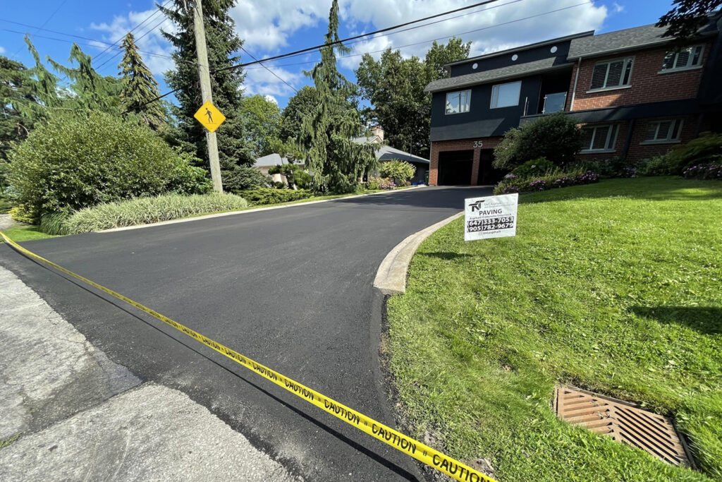 A curved driveway in front of a house that has been newly paved. There is yellow tape to block people and cars from entering, and a white sign on the lawn beside the drive with a phone number.