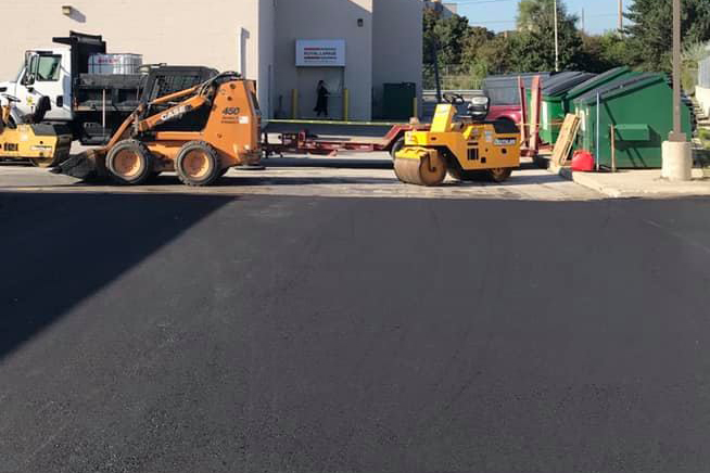 A steamroller and a bulldozer sit next to a large, newly paved parking lot behind a business.
